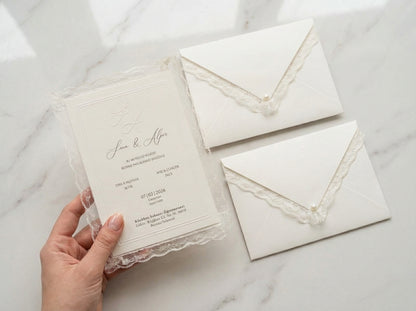 Veridia wedding invitation with delicate lace trim held over matching pearl-embellished envelopes on marble.