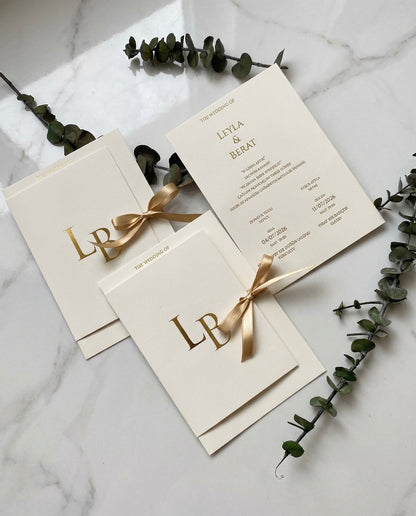 Aurielia luxury wedding invitation suite featuring gold foil monograms and silk ribbons on a white marble surface.