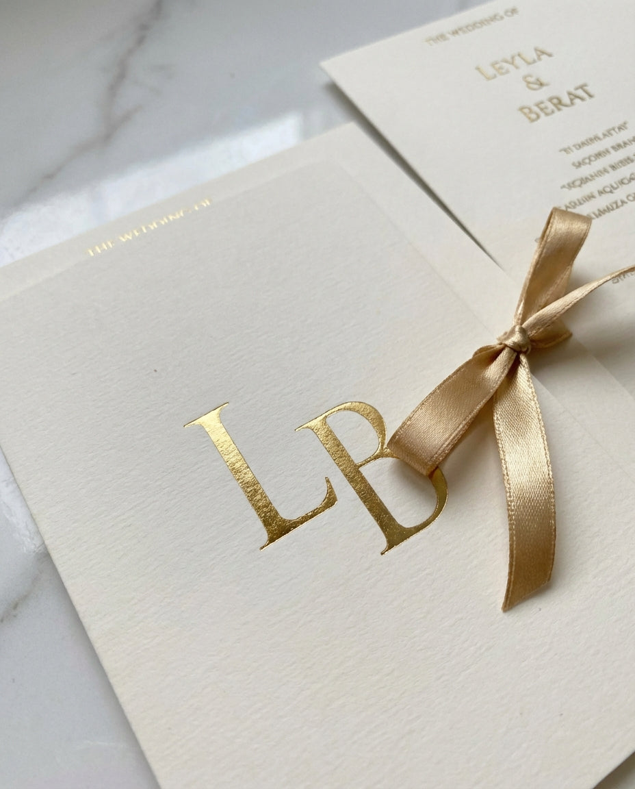 Aurielia luxury wedding invitation featuring a gold foil monogram on textured cream paper with a satin ribbon bow.