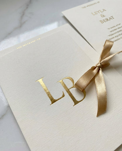 Aurielia luxury wedding invitation featuring a gold foil monogram on textured cream paper with a satin ribbon bow.